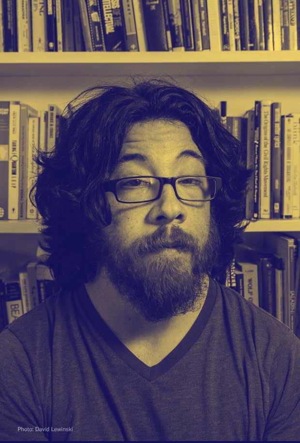 Portrait of a man with shoulder length dark hair and a beard. He is wearing plastic frame glasses and a dark t-shirt. The photo is overlaid with a yellow and blue tint. 