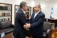 Secretary Blinken shakes hands with Prime Minister Netanyahu. There are shelves with books and an Israeli flag behind them.