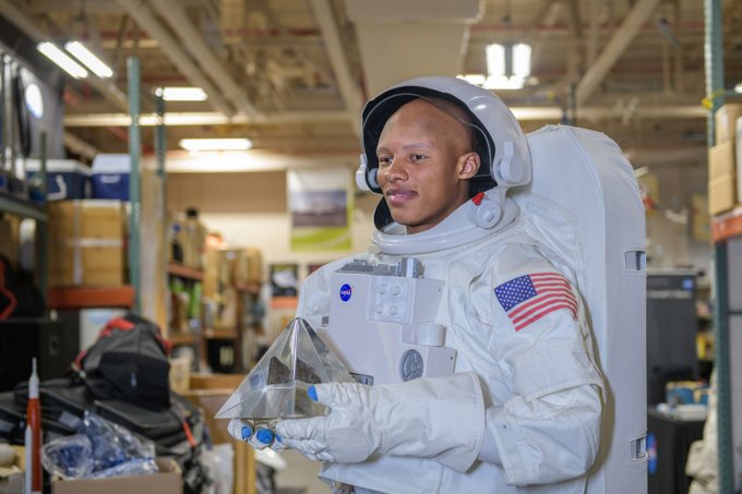 Josh Dobbs wears a white space suit and helmet replica while holding a real Moon rock. The Moon rock is enclosed in a clear, acrylic protective case. He is standing inside the exhibits warehouse at NASA's Glenn Research Center in Cleveland.