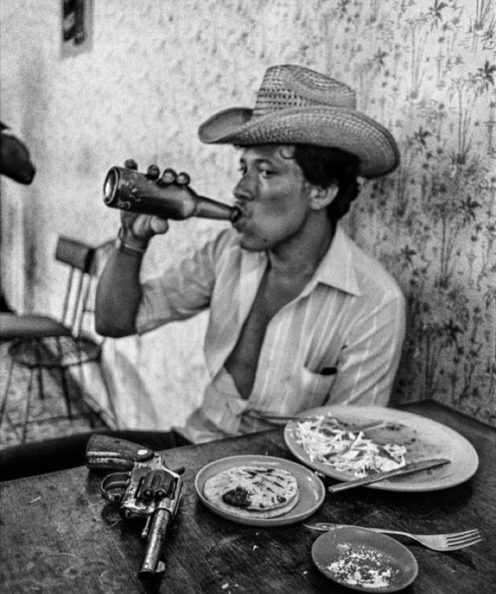 historyinmemes's tweet photo. "Death Squad" mercenary stops for lunch in El Salvador. "I bought him a beer and took three shots before he went ballistic', 1980.

This photo was taken by Derek Hudson, a well-known British photographer. Here's what he said about the incident and photo:

"I came across this guy during the civil war in El Salvador in the 1980s. He was one of a gang of mercenaries known as the Death Squad. We were in a rural backwater called Suchitoto, which at the time could best be described as a one-donkey town, and he was sitting in the local cafe having eaten his lunch, his revolver casually placed in front of him.

I bought him a beer and, as he raised the bottle, managed to get three shots in before he went ballistic. He was swearing and called his friends with rifles over and pushed me about. I don't know what would have happened if they'd realized I'd shot off some frames already. I realized I'd taken a big risk, but I didn't think about it at the time. I quickly ordered more beers, hoping to calm them down, and then just left.

I was working freelance. If you were a news photographer in New York in the 1980s, conflicts in Central America were part of the beat. I ended up going to El Salvador for weeks on end. The situation was perpetually confused - nobody really knew who was killing who. It was tense and dangerous, but there was a certain respect for the media. You felt protected in a vehicle marked "TV" or "Press," unlike nowadays in places like Syria. Of course, we did get shot at, but rarely on purpose."