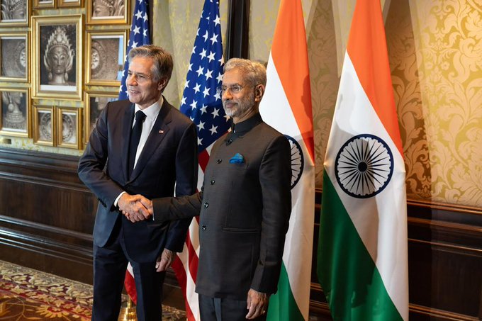 Secretary Blinken and Indian External Affairs Minister shaking hands in front of U.S. and India flags.