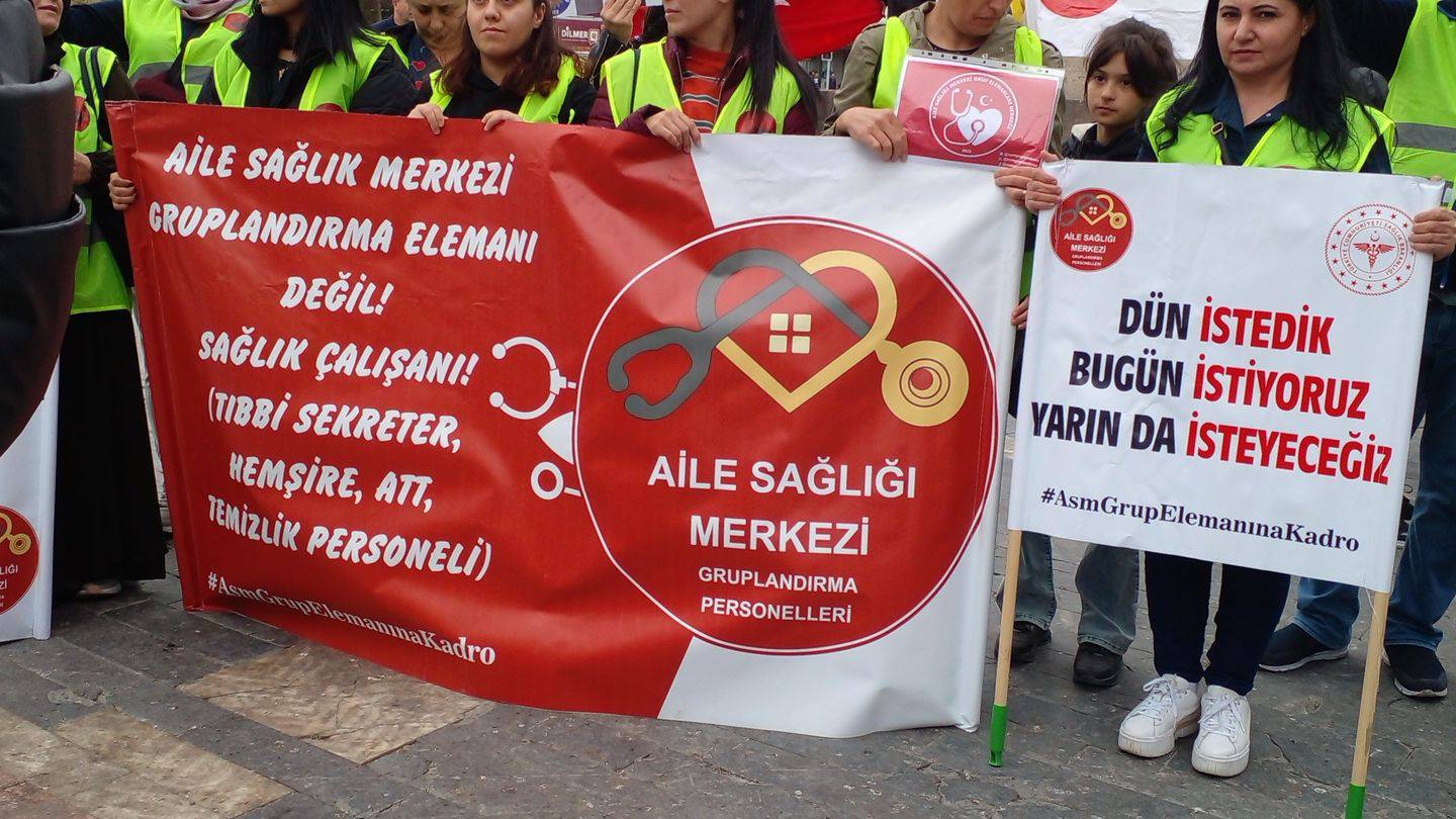 Aile Sağlığı Merkezi Gruplandırma Elemanları imzalı pankart açmış, sarı önlüklü kadın rmekçiler. Aile sağlık merkezi gruplandırma elemanı değil, sağlık çalışanı (tıbbi sekreter, hemşire, ATT, temizlik görevlisi" denilmiş pankartta