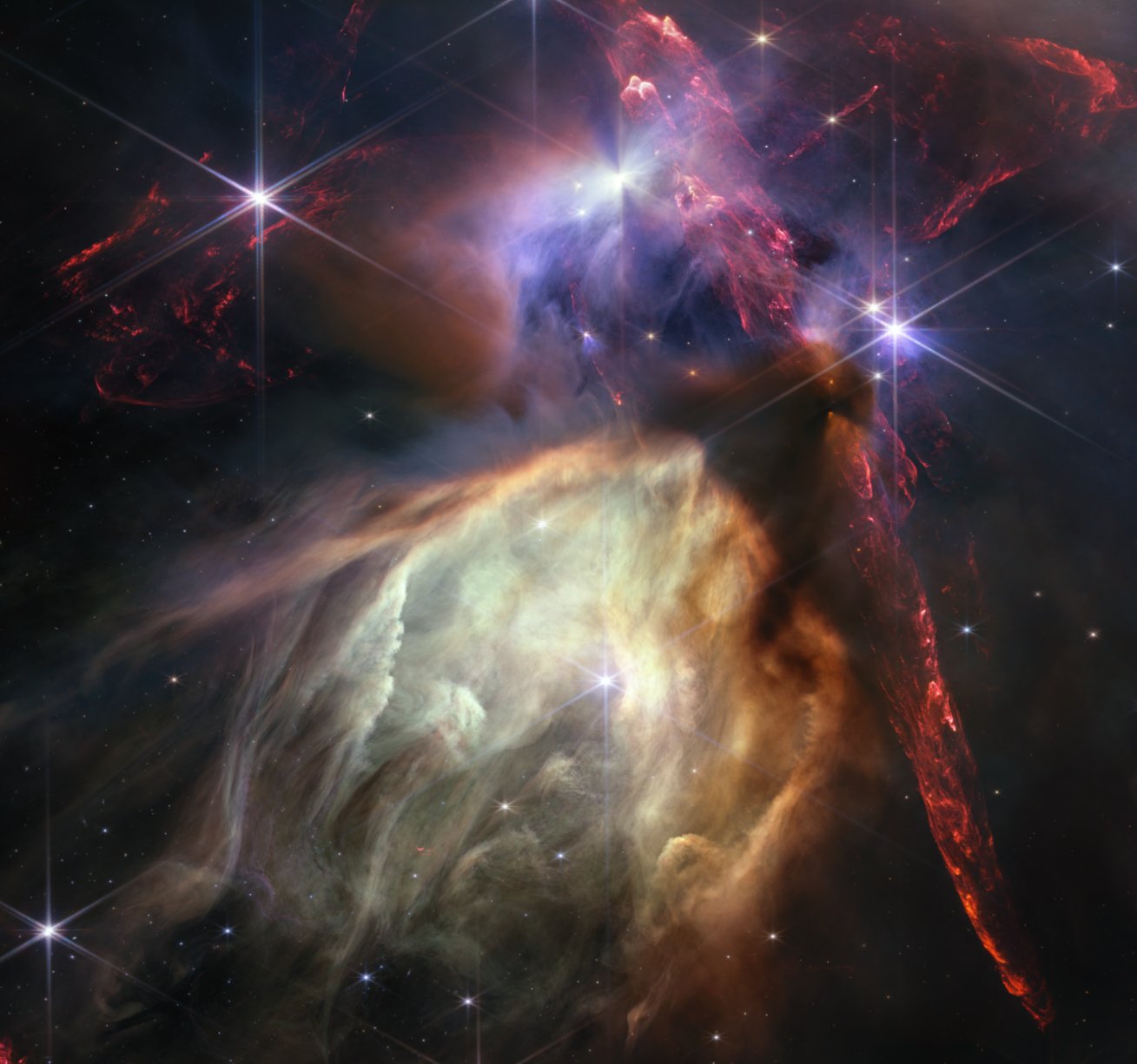 Red dual opposing jets coming from young stars fill the darker top half of the image, while a glowing pale-yellow, cave-like structure is bottom center, tilted toward two o’clock, with a bright star at its center. The dust of the cave structure becomes wispy toward eight o’clock. Above the arched top of the dust cave three groupings of stars with diffraction spikes are arranged. A dark cloud sits at the top of the arch of the glowing dust cave, with one streamer curling down the right side. The dark shadow of the cloud appears pinched in the center, with light emerging in a triangle shape above and below the pinch, revealing the presence of a star inside the dark cloud. The largest jets of red material emanate from within this dark cloud, thick and displaying structure like the rough face of a cliff, glowing brighter at the edges. Credits: NASA, ESA, CSA, STScI, Klaus Pontoppidan (STScI), Image Processing: Alyssa Pagan (STScI)