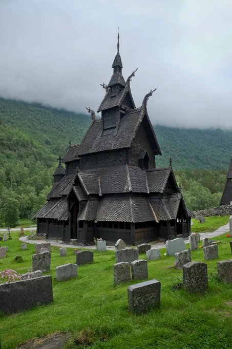 a weathered wooden church with many gables sitting among graves on a lush green hill, in front of a foggy forested landscape