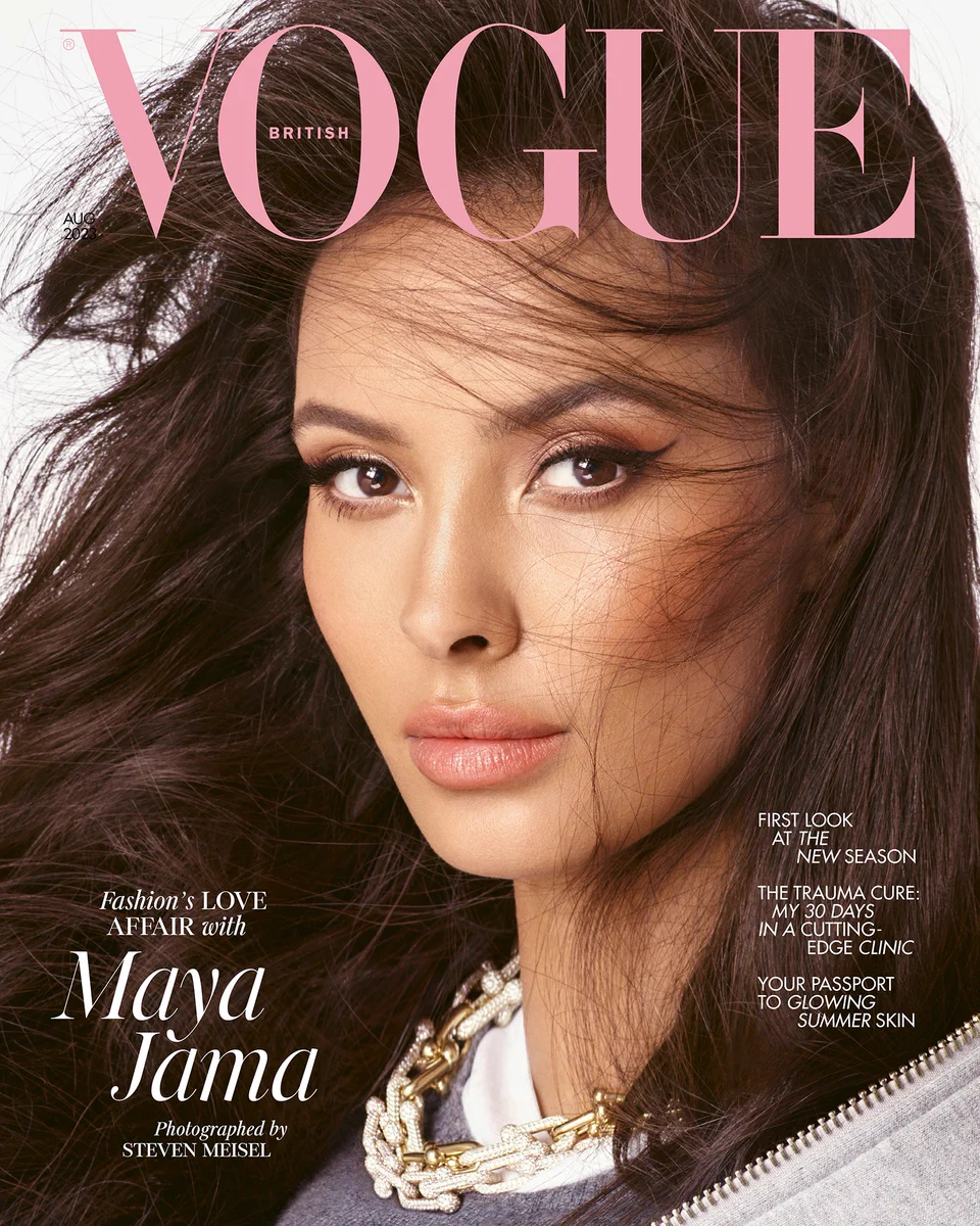 Image is a close-up shot of Maya Jama, a mixed-race woman with windswept dark brown hair, on the cover of British Vogue. She wears winged liner and a sweep of bronzer with a peachy lip. Just visible in the bottom of the image are her grey hoodie sweater and T-shirt, both by Miu Miu, worn with a gold and diamond link necklace by Tiffany & Co. Above her head, the Vogue logo appears in coral pink, while a cover line declares: “Fashion’s love affair with Maya Jama.”