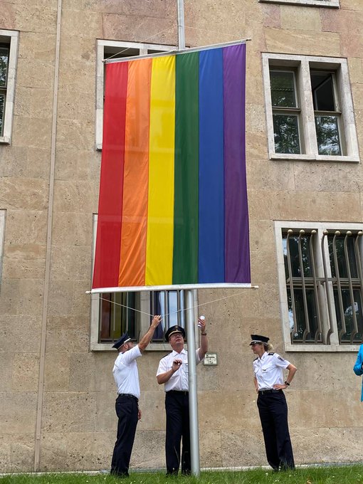 Drei Personen in Uniform stehen an der Regenbogenflagge.