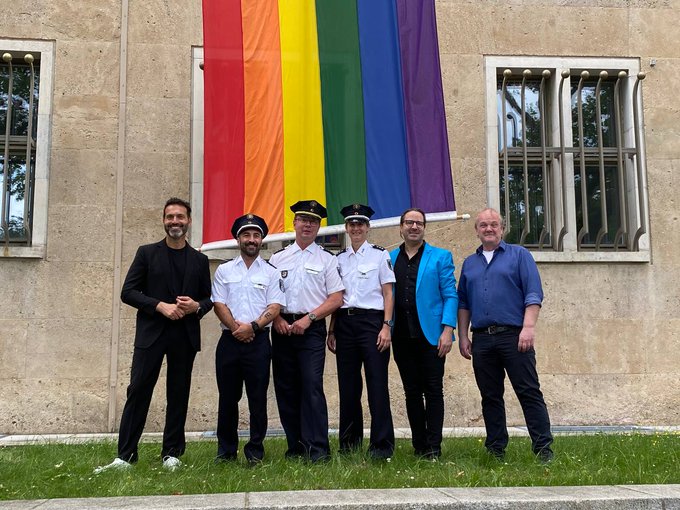 Fünf Personen, darunter drei in Uniform, stehen vor der gehissten Regenbogenflagge.