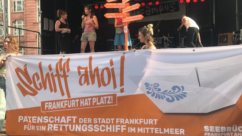 Vor einer Bühne auf dem Campus Bockenheim halten Menschen ein orange-weißes Transpi hoch: SCHIFF AHOI - Frankfurt hat Platz!<br>Auf der Bühne zwei Personen im Gespräch. Eine weitere Person hält einen orangen Pfahl mit orangenen Wegweisern: Flughafen Frankfurt / Warschau, Hauptsitz von Frontex / Abschiebegefängnis DA-Eberstadt / Grenze Polen-Belarus / Mittelmeer / Berlin, Verkehrsministerium.