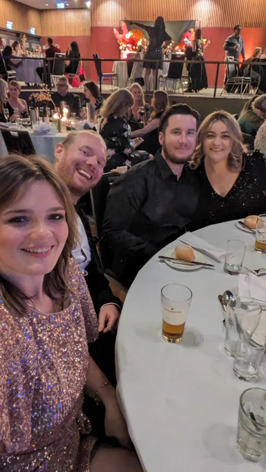 Four smiling people dressed in formalwear seated at a table with a white tablecloth. Behind them is a room full of other tables.