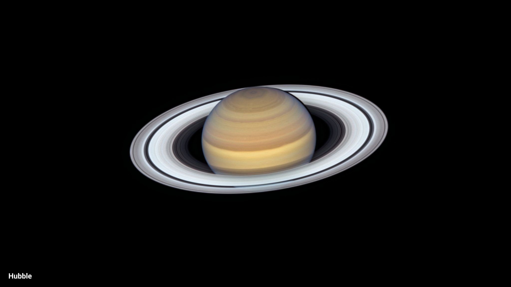 Against the black background of space, the planet Saturn glows in bands of yellow and brown. It's surrounded by pale rings, seen almost face-on.
