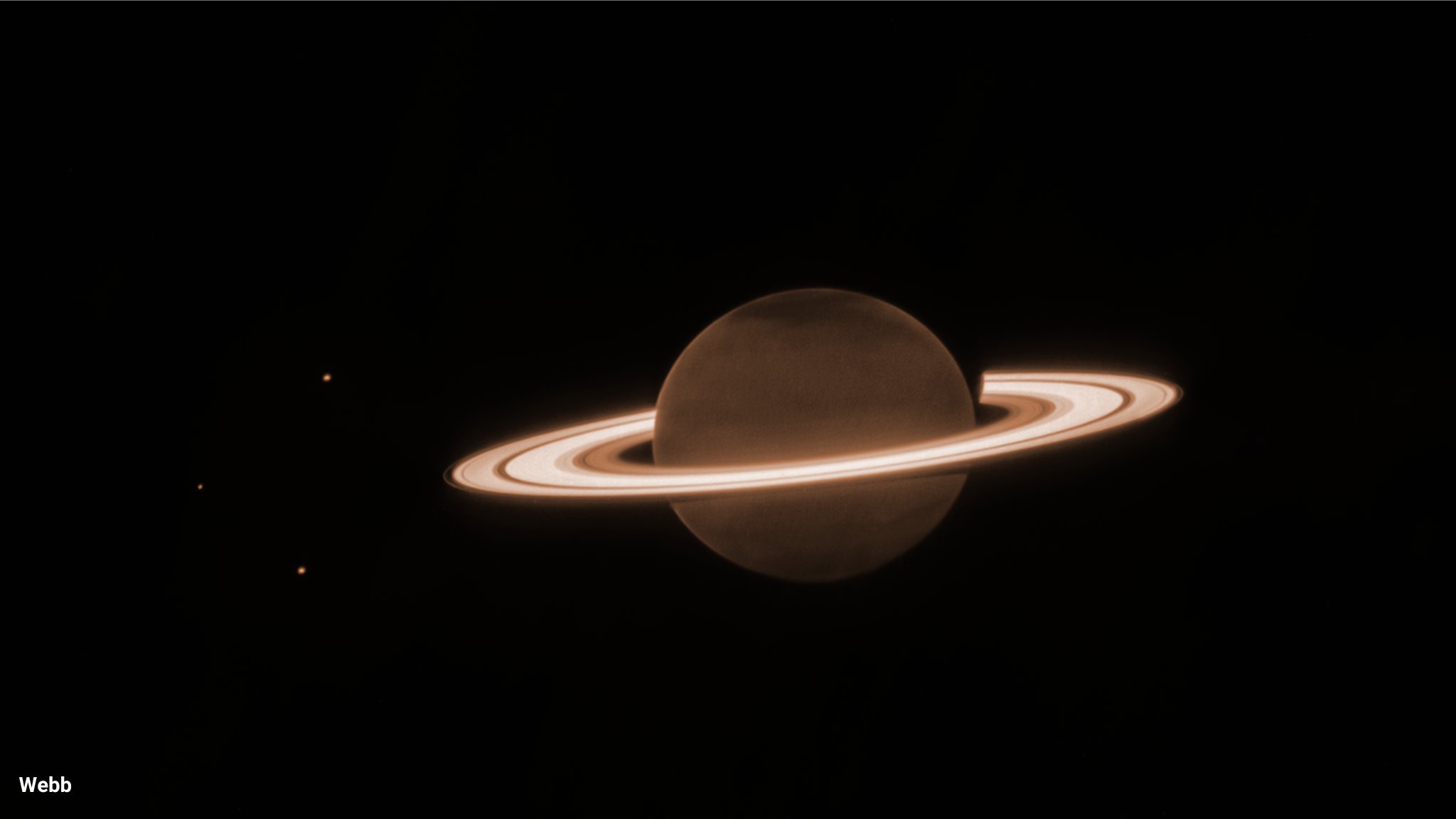 The background is mostly dark. At the center is a dark orange-brown circle, surrounded by several blazing bright, thick, horizontal whiteish rings. This is Saturn and its rings. There are three tiny dots in the image—one to the upper left of the planet, one to the direct left of the planet, and the lower left of the planet. These are three of Saturn’s moons: Dione, Enceladus, and Tethys, respectively. There is a slightly darker tint at the northern and southern poles of the planet. The rings surrounding Saturn are mostly broad, with a few singular narrow gaps between the broader rings. There is an innermost, thicker ring, and next to that is a brighter, wider ring. Traveling farther outward, there is a small dark gap before another thicker ring. In the thicker ring, there is a narrow faint band. There is then an outermost, faintest, thinnest ring.