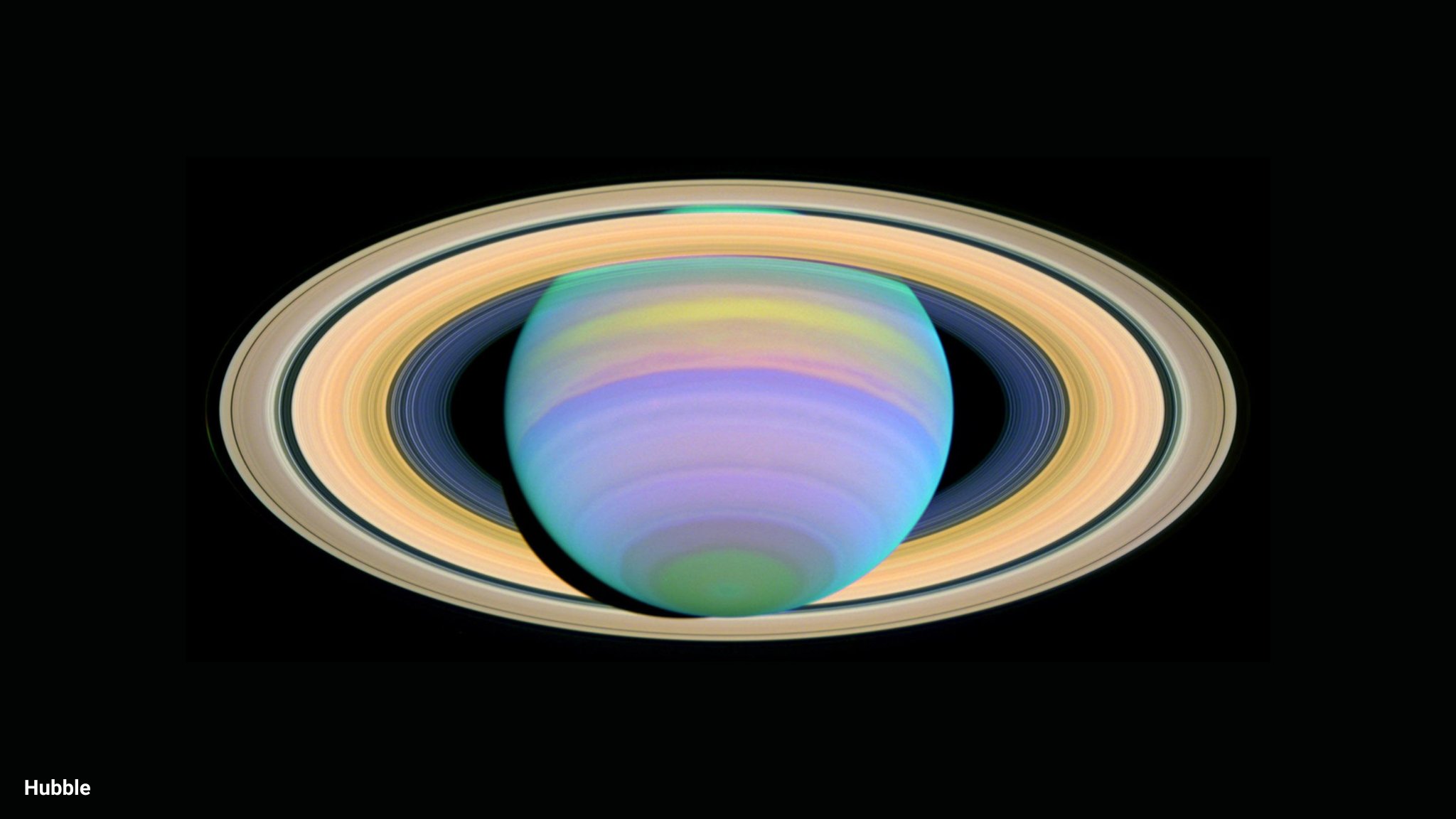 Against the black background of space, the planet Saturn glows in colorful bands of purple, green, yellow, and pink. Its rings are seen almost face-on in a yellowish hue. 