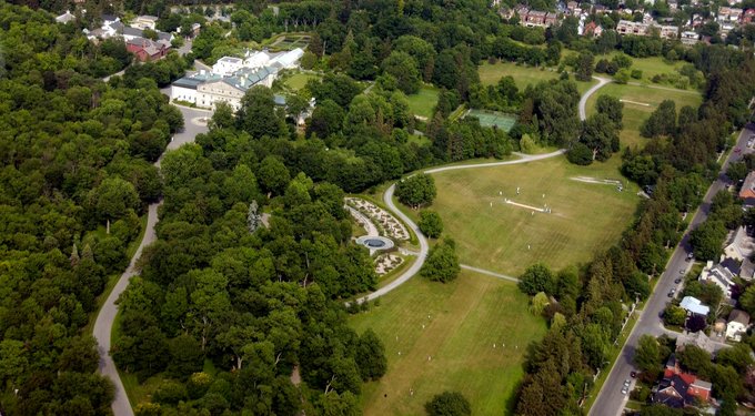  An aerial view of the grounds of Rideau Hall. On the right, there is a cricket field with players; on the left, there is a tree-lined pathway leading to a building. There is a rose garden located between the two.