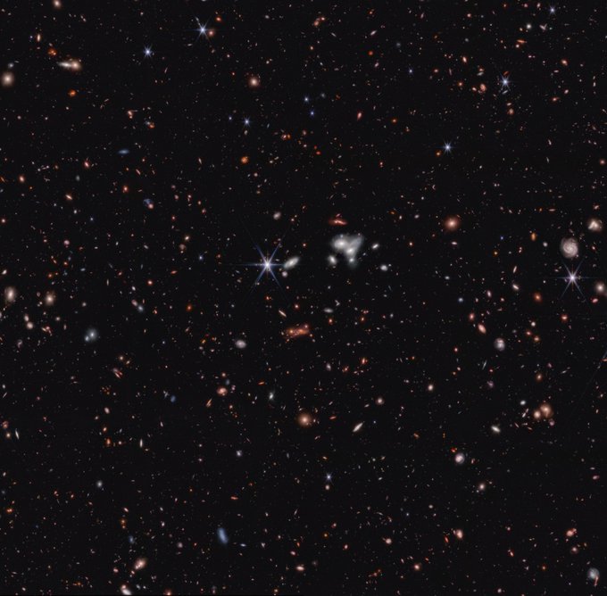 Thousands of galaxies appear in this view, which is set against the black background of space. There are many overlapping objects at various distances. They include large, blue foreground stars, some with all eight diffraction spikes, and white and pink spiral and elliptical galaxies. Numerous tiny red dots appear throughout the scene. This is a portion of a vast survey known shorthand as CEERS.