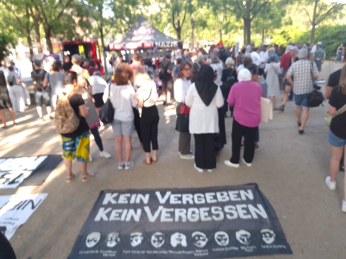 Menschen auf dem Dernschen Gelände in Wiesbaden um einen Lauti versammelt. Im Vordergrund ein Transpi KEIN VERGEBEN - KEIN VERGESSEN mit Porträts der 9 am 19. Februar ermordeten jungen Menschen.