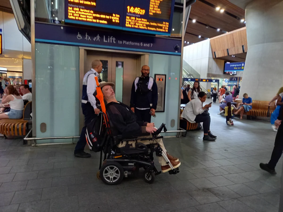 A lift on a station concourse, above which is an orange lit displayboard. Standing around are a group of staff in hi-viz jackets and tabbards. With them is Alan in his wheelchair wearing a dark coat.