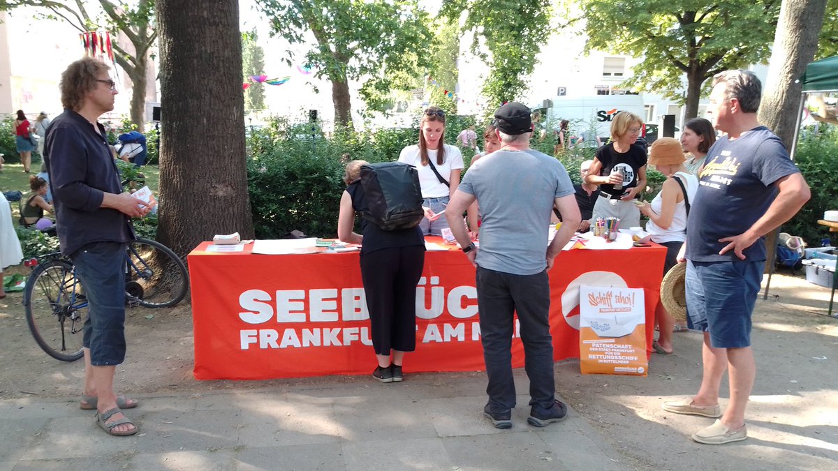 Oranger Seebrücke Infostand auf dem Kurfürstenplatz mit Menschen davor, die sich informieren. Eine Person unterschreibt.