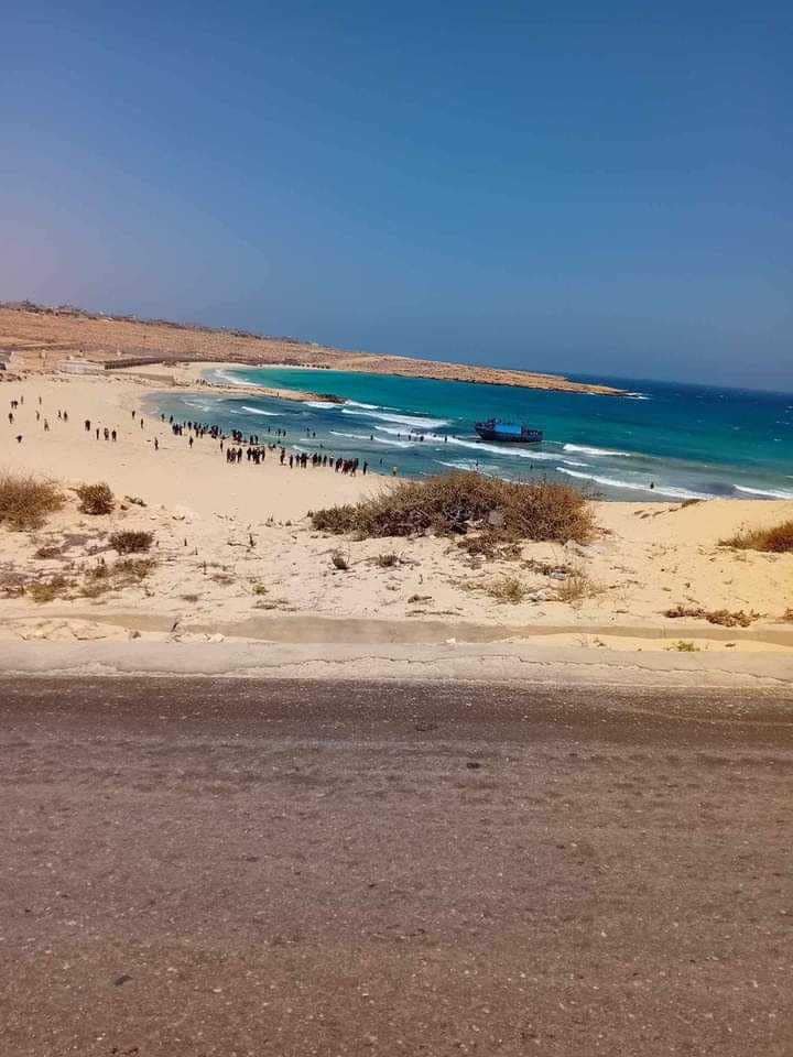 Das nach Libyen zurückgekehrte Boot ist an einem Strand auf Grund gelaufen. Menschen waten durch das Wasser ans rettende Ufer, wohin zum Zeitpunkt der Aufnahme knapp hundert Menschen am Ufer stehe.