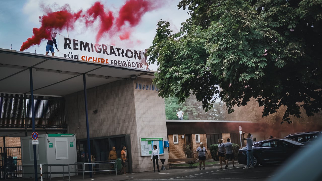 Aktionsfoto mit Banner und rotweißem Rauch. Auf dem Banner steht "Remigration für sichere Freibäder"