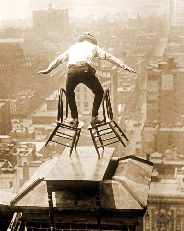 A daredevil balances on the roof edge of a Manhattan building, perched on a table placed over a chimney, 1910s.