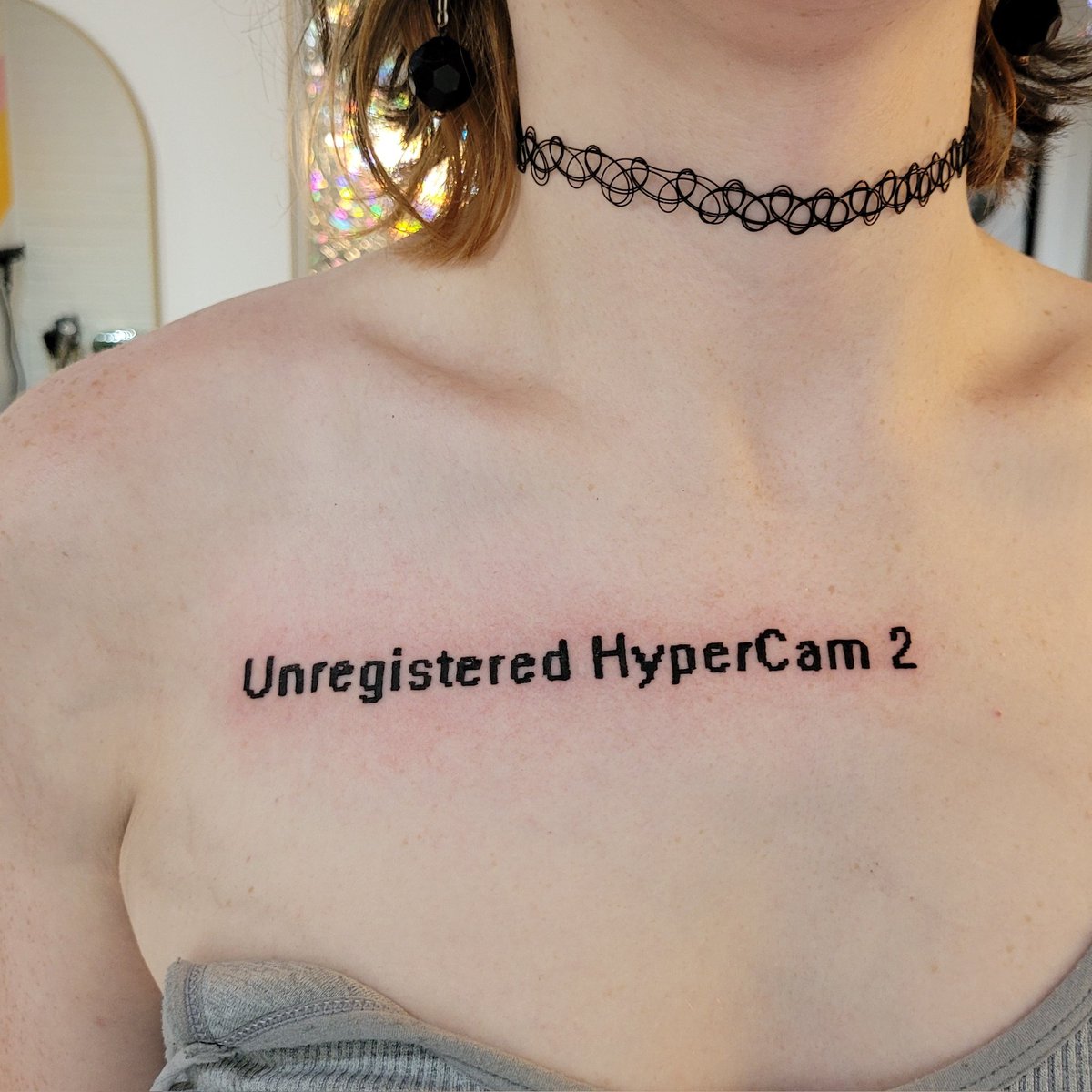 a fresh tattoo on the upper left chest, below the collarbone of a person. it is a pixel text tattoo that reads, “Unregistered Hypercam 2” in reference to a watermark displayed in the upper-left corner of footage recorded with free versions of the HyperCam 2 screen capture software developed by Hyperionics, Inc. this program was used in the early days of youtube for let’s plays, tutorials and other screen recording needs.