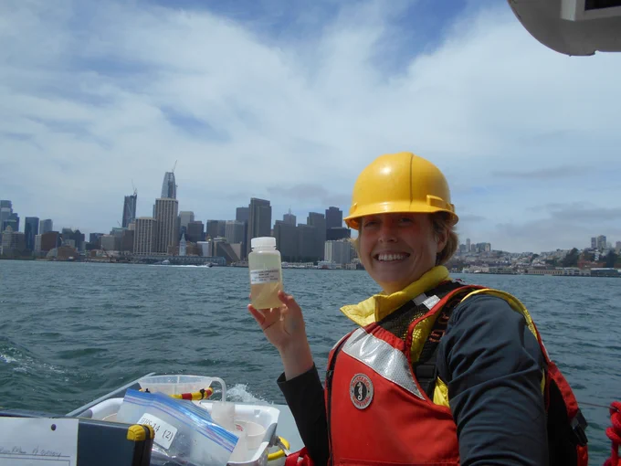 Young woman wearing a red life jacket and yellow hard hat sits on a boat, holding up a clear plastic container with a water sample. The city shoreline stretches out in the background behind her.