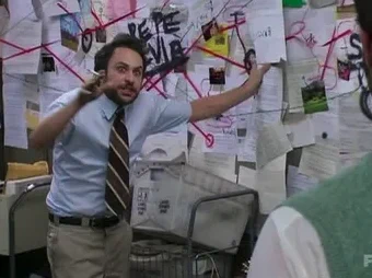 frantic man stands in front of cork board filled with papers with a wild look in his eyes
