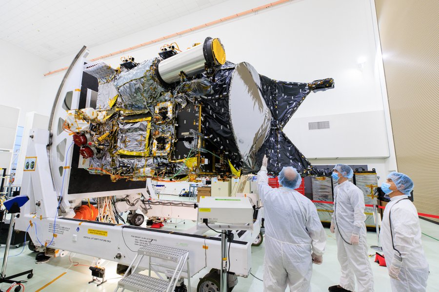 NASA’s Psyche spacecraft is shown in a clean room on June 26 at the Astrotech Space Operations facility near the agency’s Kennedy Space Center in Florida. The spacecraft is boxy and about the size of a car. From this side view many wires are visible, however, much of the surface of the spacecraft is covered by black, gold, and silver protective blankets. Hundreds of small two-inch sized pieces of silver and gold tape hold the wire and blankets into place. Two holes can be seen on the side where an array made of four solar panels will later be attached. Three team members stand to the right of the spacecraft wearing protective white uniforms, latex gloves, face masks, and hair nets. They are looking up at the spacecraft as one of the team members points to it. Credit: NASA/Frank Michaux