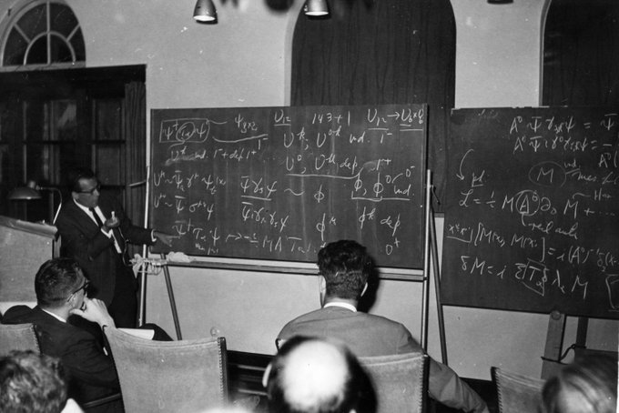 Julian Schwinger lecturing a class. He is seen pointing toward what seems to be a quantum mechanical function written on the chalkboard.
