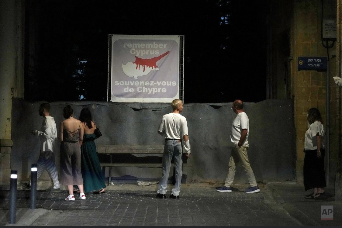 pkaradjias's tweet photo. Tourists stand by a wall blocking a road with barrels and sand bags, with a banner showing the island in division, across the UN buffer zone that divides the Greek, south, and Turkish Cypriots, north, areas, in Nicosia, Cyprus, Thursday, July 20, 2023. (AP Photo/Petros Karadjias) https://t.co/9rigt78wsM