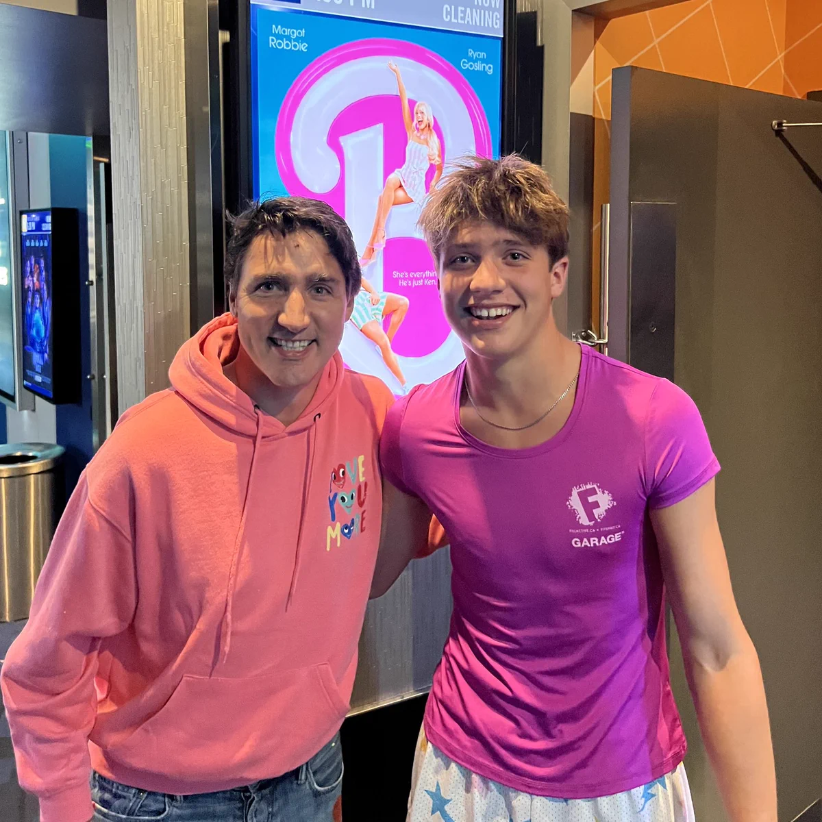 Prime Minister Justin Trudeau and Xavier Trudeau stand in front of a movie theatre entrance. They are dressed in pink. Behind them is a sign for the Barbie movie.