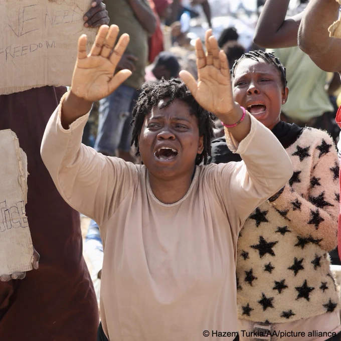 Two women crying. Next to them is a person holding up signs, asking for help.