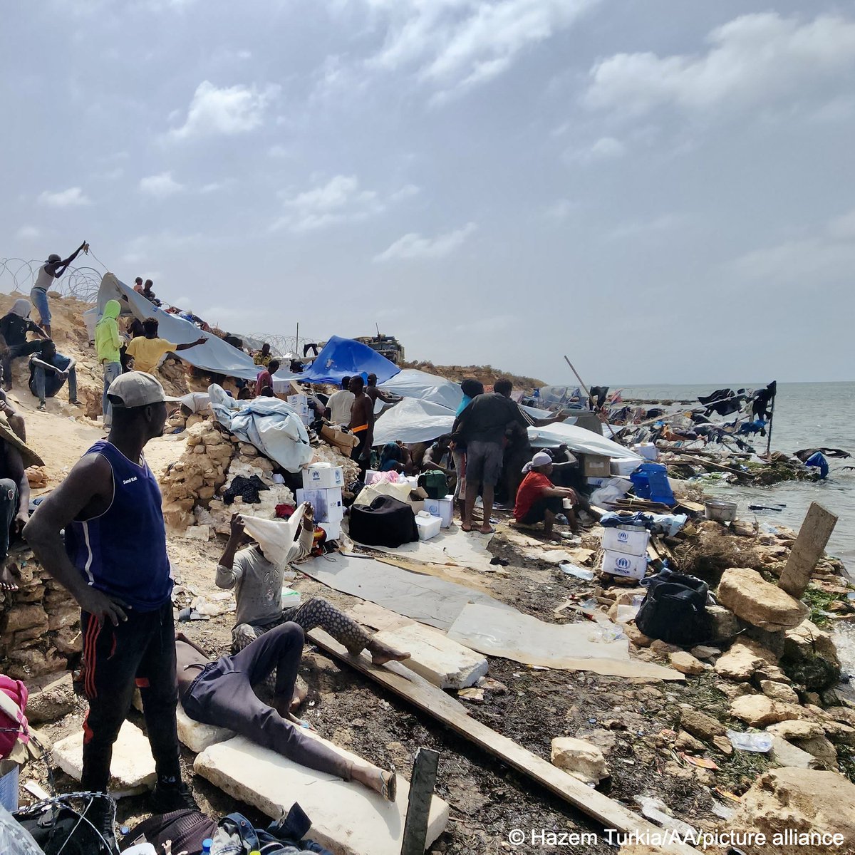InfoMigrants's tweet photo. 📷 These migrants are stuck at the Tunisian-Libyan border. 

They were forced into the border region by Tunisian forces, and Libyan border guards reportedly won’t let them cross. 

Now, they are living in a makeshift camp in Ras Ajdir next to the barbed-wire border fence. https://t.co/hzmcr6GUfs