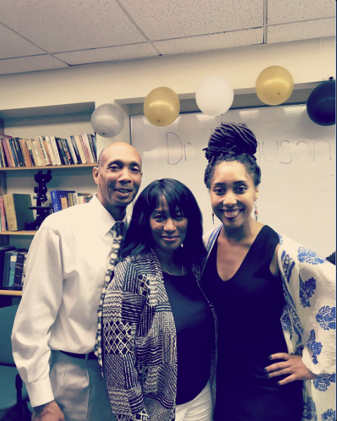 Brielle is standing in a conference room with her parents smiling wide after her PhD defense. A white board behind her is decorated with balloons and reads Dr. Ferguson. Brielle's hair is in faux locs, and in a high bun. 