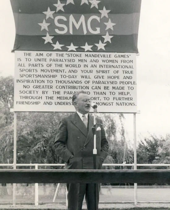 Sir Ludwig Guttmann stands in front of a sign for the Stoke Mandeville Games. The sign reads: The aim of the Stoke Mandeville Games is to unite paralysed men and women from all parts of the world in an international sports movement, and your spirit of true sportsmanship today will give hope and inspiration to thousands of paralysed people. No greater contribution can be made to society by the paralysed than to help, through the medium of sport, to further friendship and understanding amongst nations.