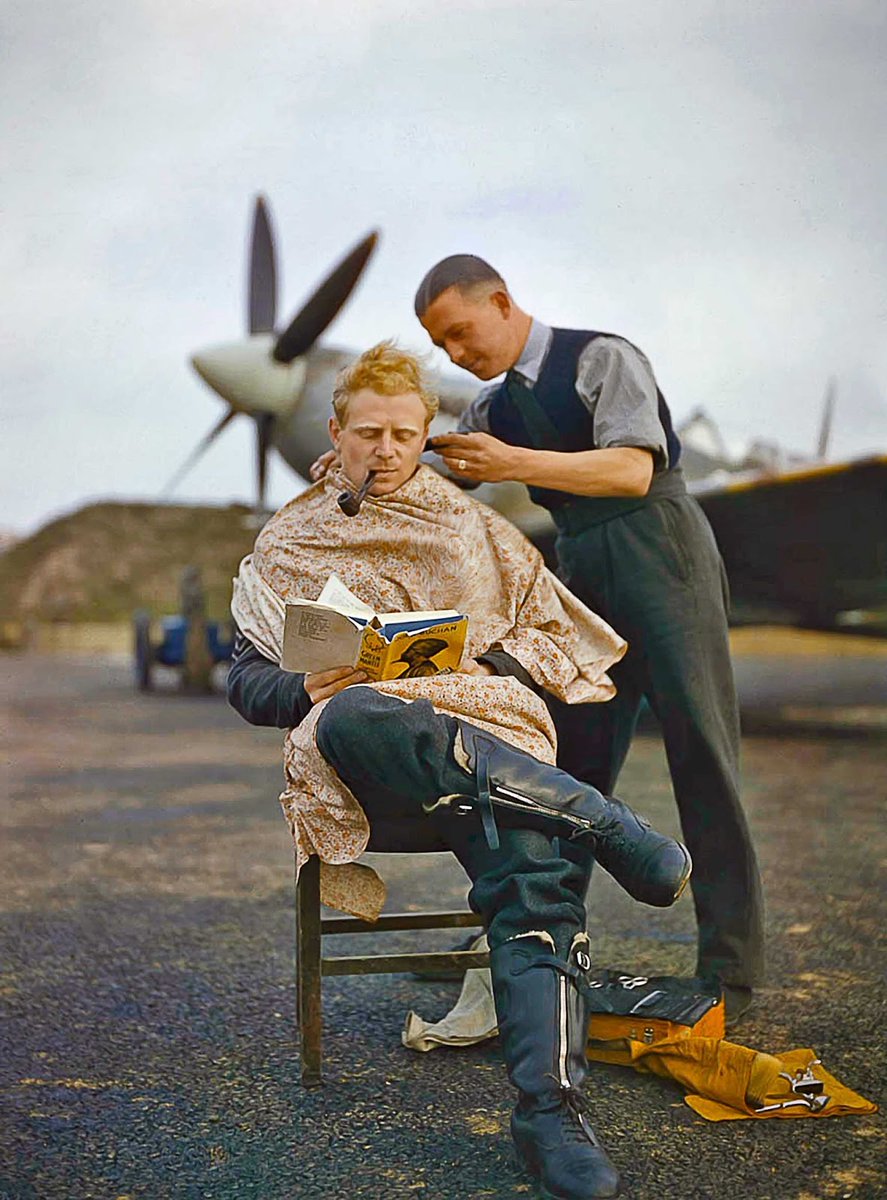 This striking image shows a British Royal Air Force pilot receiving a haircut while leisurely reading a book and smoking his pipe, all while dressed for battle with his spitfire ready to take off at a moments notice in the background.

The calmness of the man is surprising and at
