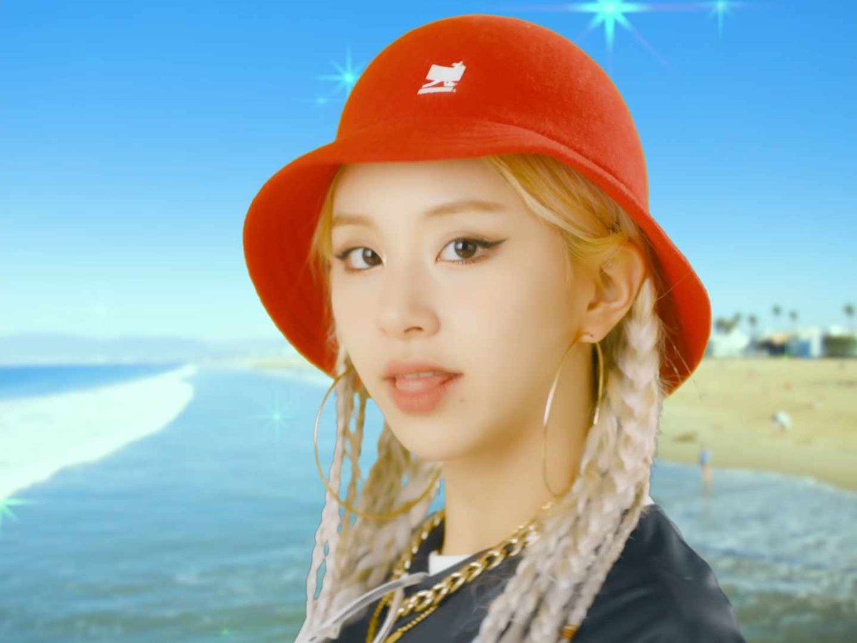 A portrait of a young woman with blonde braided hair wearing a red bucket hat and large gold hoop earrings, set against a blurred beach background with sparkles added to the image.