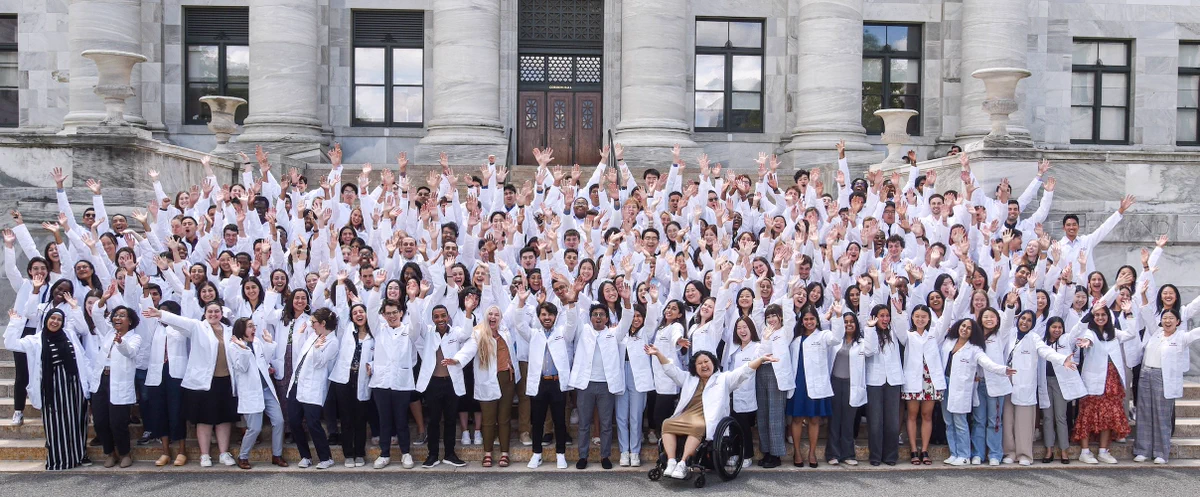 A large group of students in white coats stand on the steps of Gordon Hall waving their hands in the air