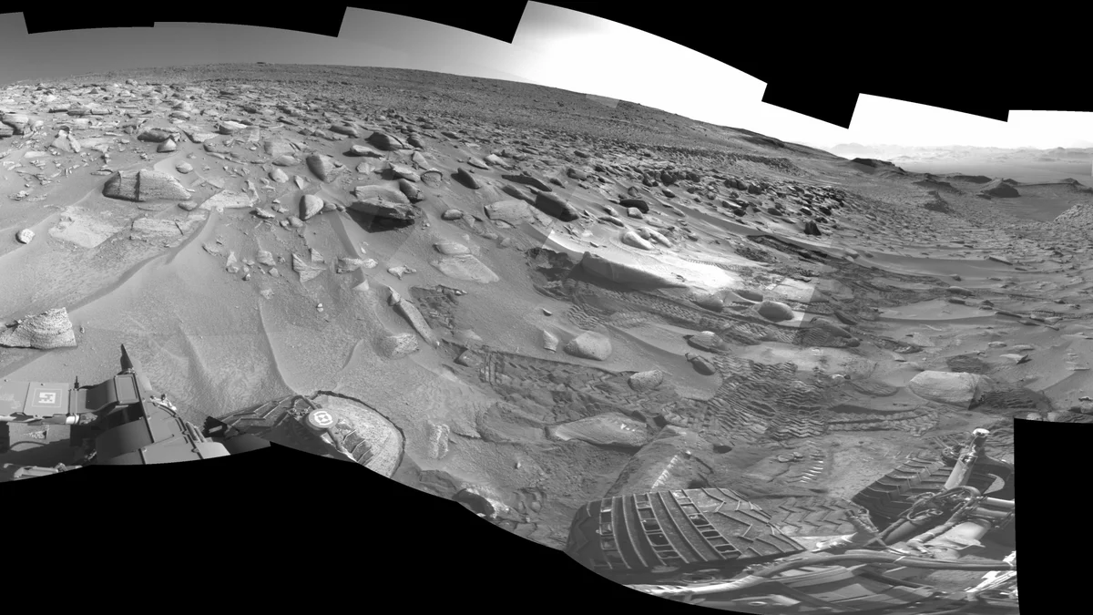 A series of images from the Curiosity rover captured the slippery terrain it faced during a recent climb up Mount Sharp on Mars. Along the top and bottom are boxy cutouts of black where images weren't taken. The rest of the scenery is black and white and shows parts of the rover's wheels and front part of its body. The landscape is covered in large rocks and shows sandy terrain and dozens of rover tracks along the right side that indicate the slippery surface.