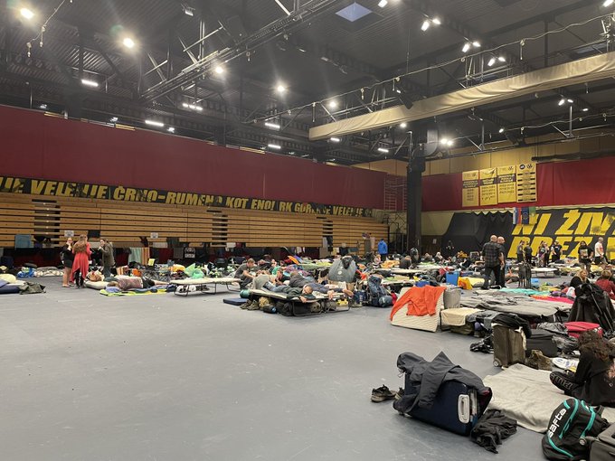 Foto van een sporthal waarin allerlei matjes en campingspullen staan. Mensen slapen, vermaken zich met spelletjes.