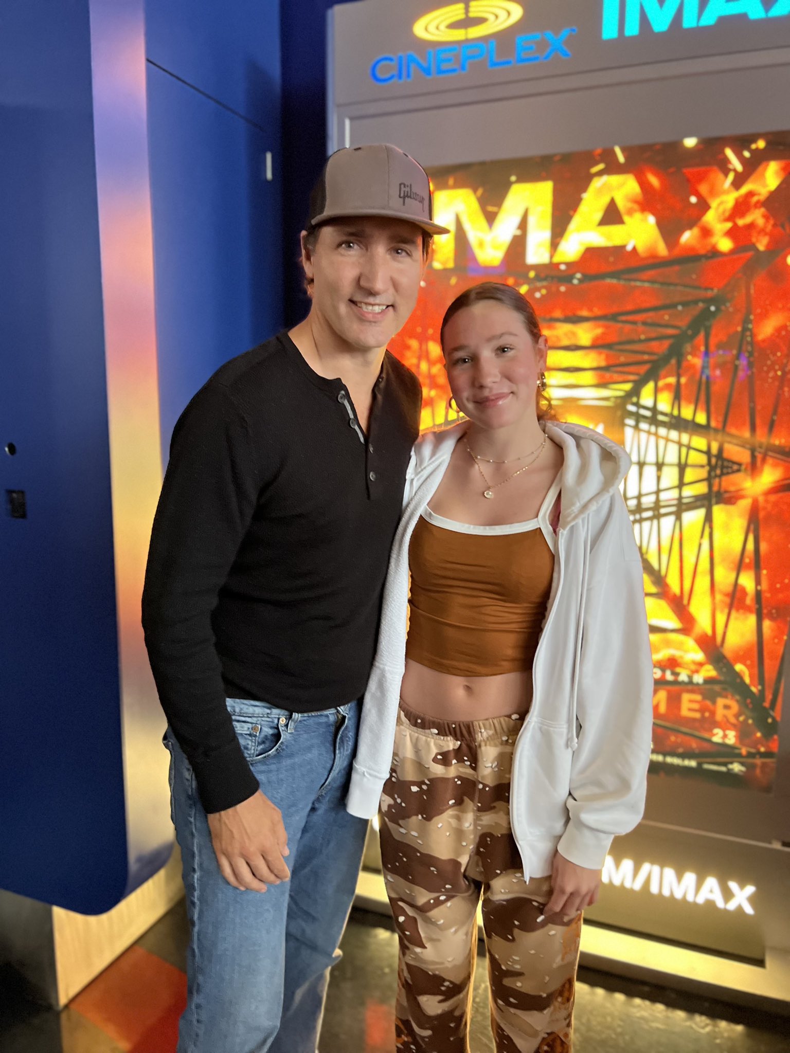 Prime Minister Justin Trudeau and Ella-Grace Trudeau stand beside each other in front of a movie theatre entrance. Behind them is a sign for the Oppenheimer movie.
