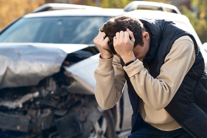 Distressed young man clutching his head with a crashed car behind him.