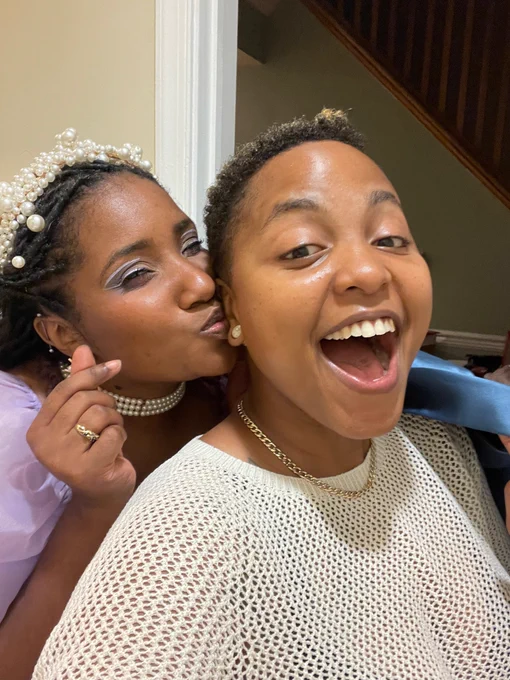 I’m in a purple dress with pearls on my neck and crown and earrings. I’m making a little heart with my pointer finger and thumb. I’m kissing my partner’s cheek. My partner has pearl gauges and gold chain and a tan mesh shirt. She is also opening her mouth. we are two black women.