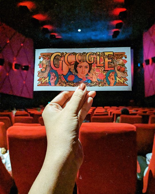 An image taken inside a movie theatre, with a person's hand holding a piece of paper with the Google Doodle for Sridevi printed on it. The paper and the movie screen are aligned with each other.