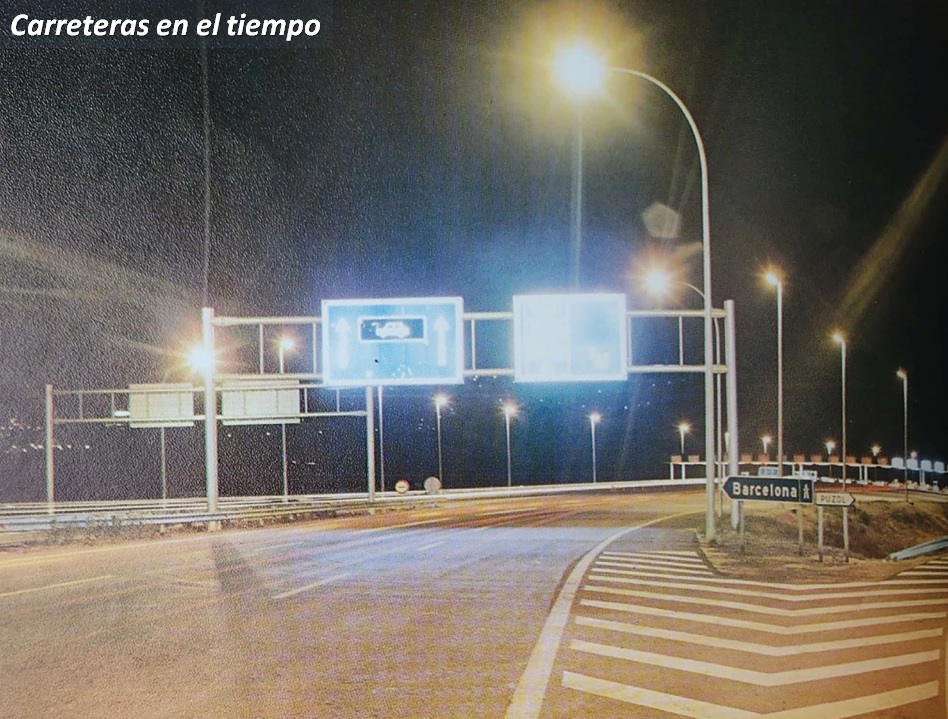 Vista nocturna de la autopista del Mediterráneo, actual AP-7 en las inmediaciones de la antigua estación de peaje trocal de Puzol. En ella destaca la singular señalización vertical elevada en pórticos compuesta por carteles con iluminación interior. Fuente de la fotografía: AUMAR