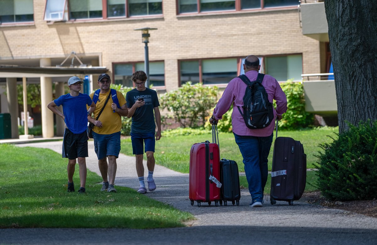 A student in a purple shirt wheels three suitcases of various sizes down a concrete path, while three people walk past.