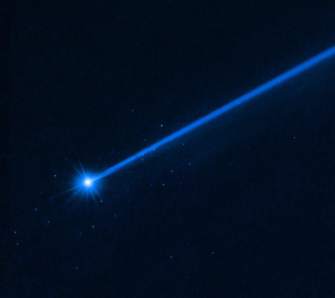 The bright white object at lower left is the asteroid Dimorphos. It has a blue dust tail extending diagonally to the upper right. A cluster of blue dots surrounds the asteroid. These are boulders that were knocked off the asteroid.