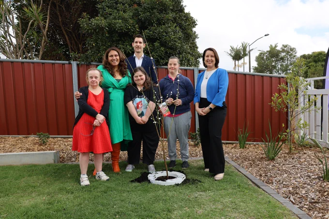 (Left to right) Susie, Angie and Alison are standing in a group with three other people in a yard area. Susie is wearing a red dress and dark long-sleeved cardigan, Angie is wearing a dark shirt with the cartoon mermaid Ariel printed on it and dark pants, Alison is wearing a blue jumper and grey pants. The woman next to Susie is wearing a green dress, the man behind the group is wearing a blue suit and the woman to the right of Alison is wearing a light blue jumper, a white shirt and dark pants. Everyone is smiling for the camera and there is a tall plant in front of the group.