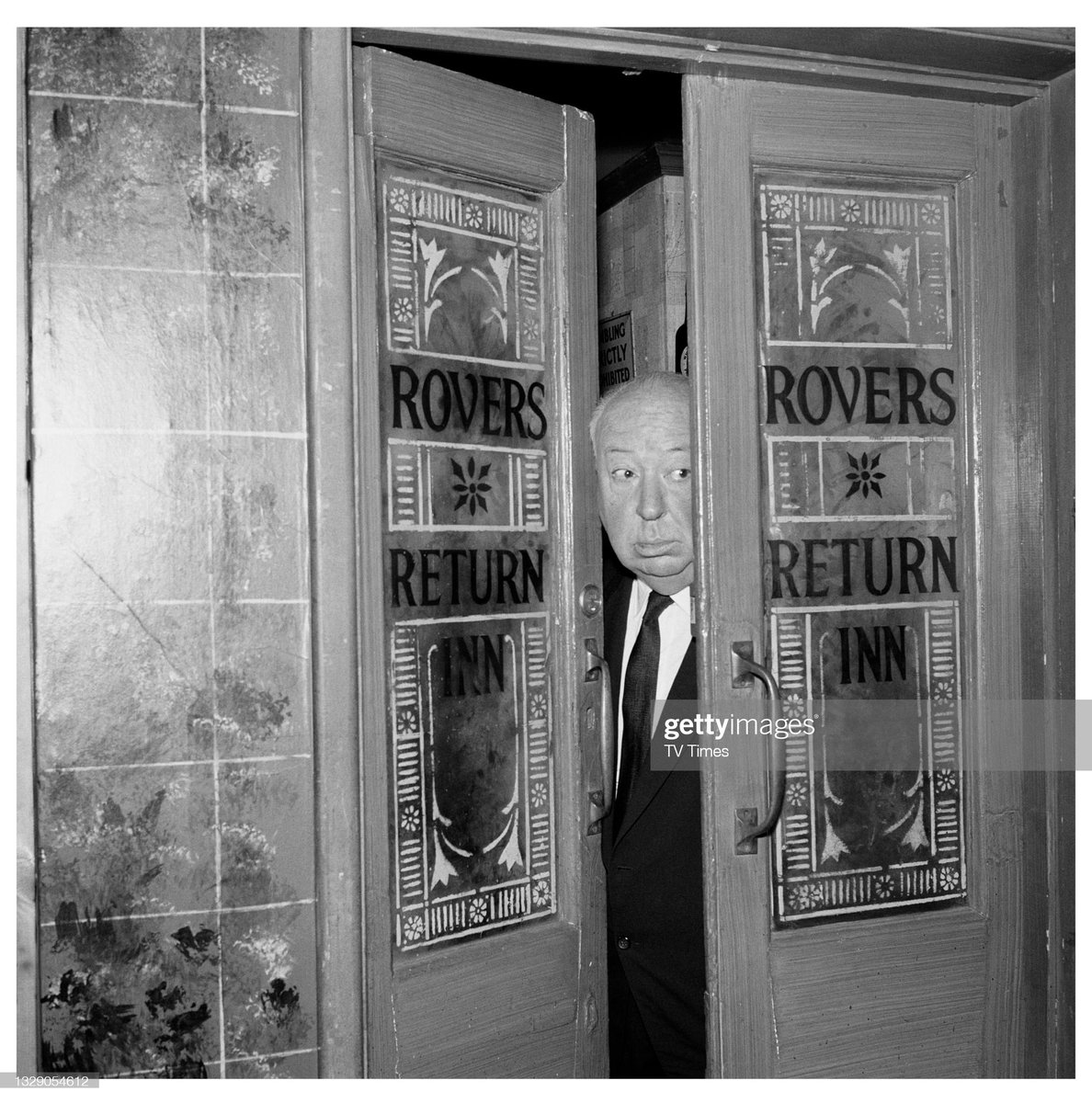 English film director Alfred Hitchcock photographed poking his head through the doors of the Rovers Return Inn on the set of television soap Coronation Street, at Granada Studios in Manchester (1964)