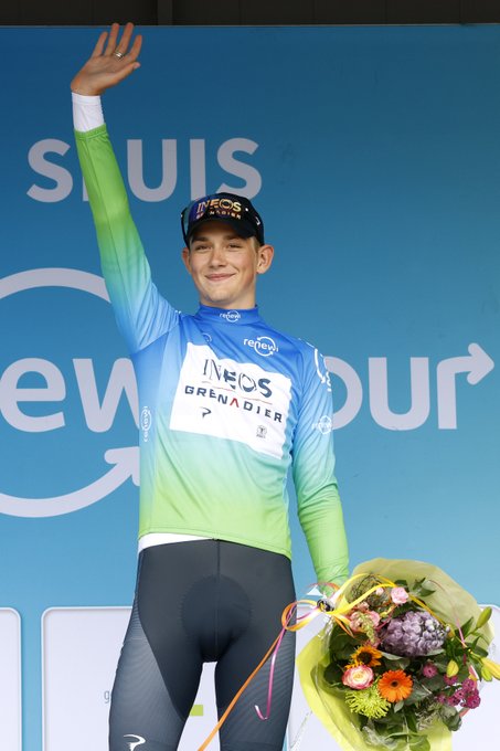 Josh Tarling in a blue and green leaders jersey holding a bunch of flowers.